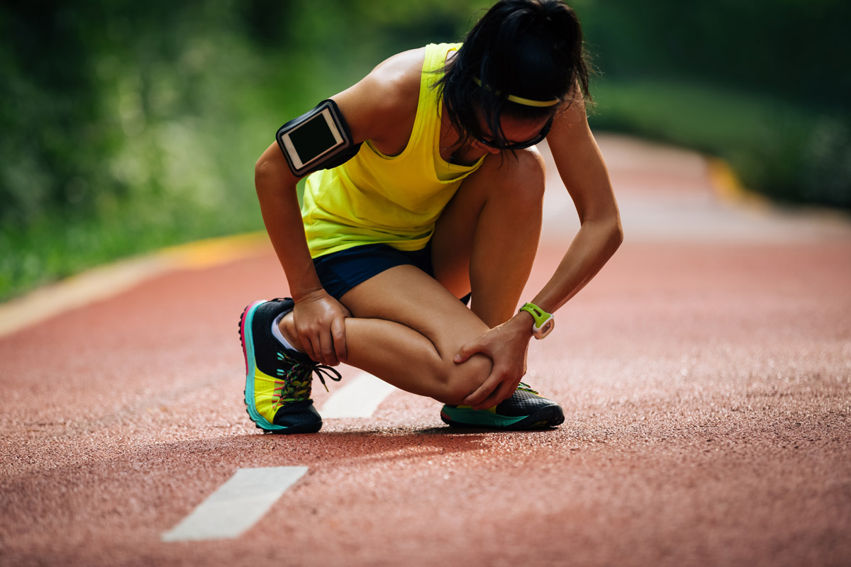 athletic woman bent over holding shin in pain while on a run outdoors