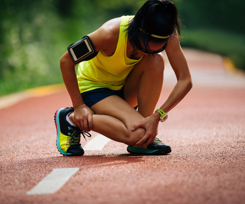 athletic woman bent over holding shin in pain while on a run outdoors