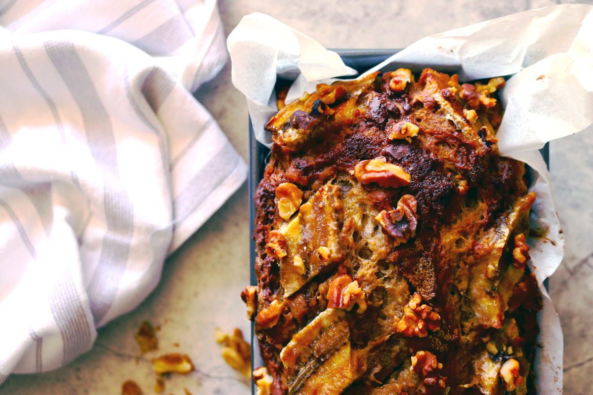 a freshly baked loaf of Banana Walnut Bread in a baking pan cools on a marble counter with a striped dish towel and walnut pieces surrounding the loaf