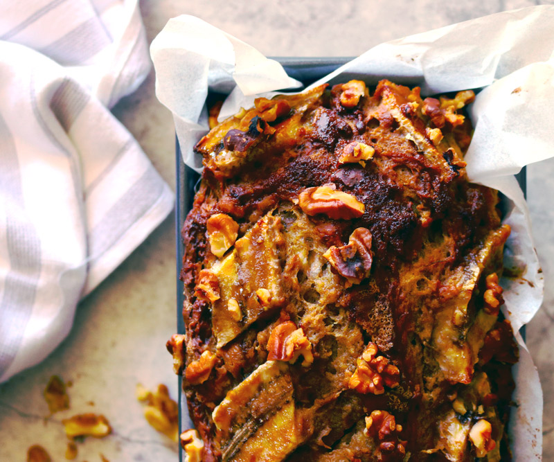 a freshly baked loaf of Banana Walnut Bread in a baking pan cools on a marble counter with a striped dish towel and walnut pieces surrounding the loaf