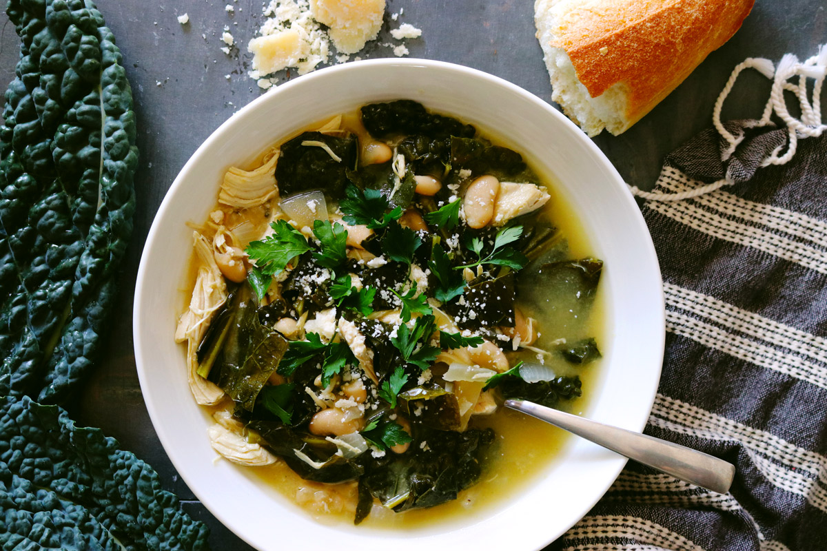 a bowl of Slow Cooker Chicken Stew surrounded by kale leaves, crumbled Parmesan cheese and a torn baguette