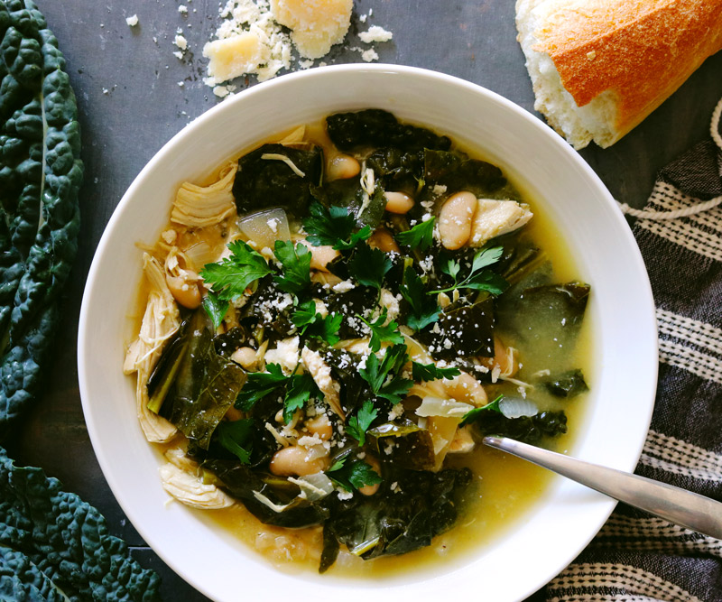 a bowl of Slow Cooker Chicken Stew surrounded by kale leaves, crumbled Parmesan cheese and a torn baguette