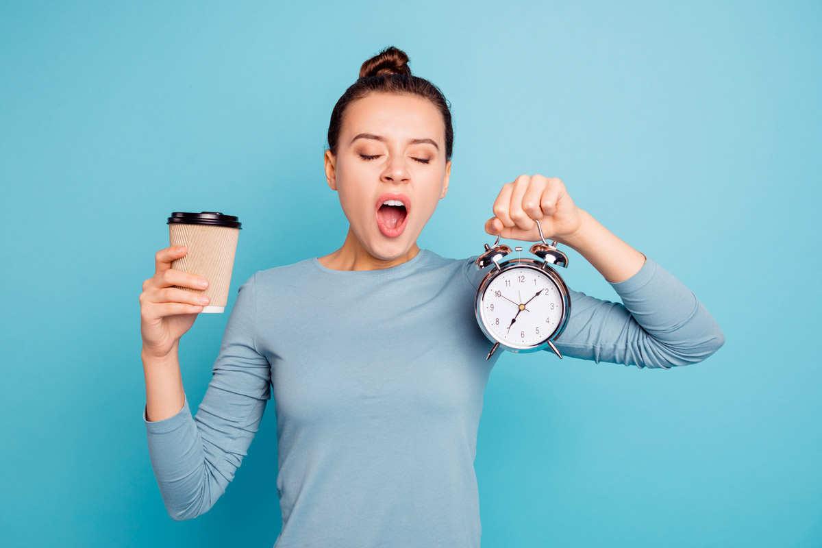 woman yawning holding an alarm clock in one hand and a cup of coffee in the other hand