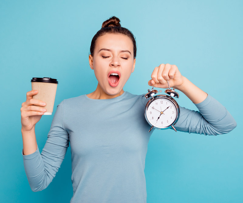 woman yawning holding an alarm clock in one hand and a cup of coffee in the other hand