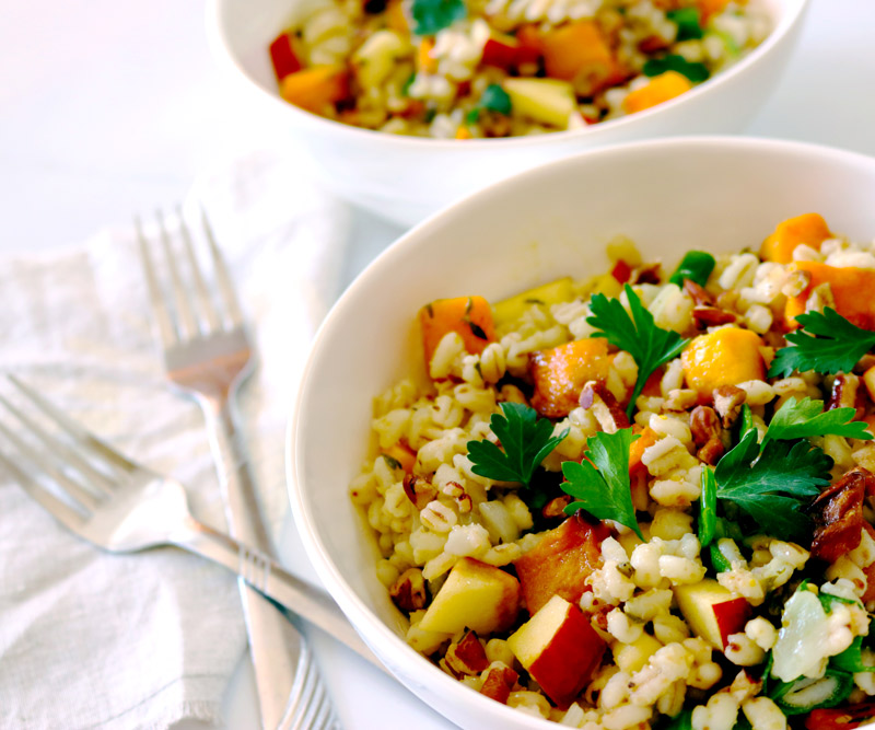 two white bowls filled with Fall Farro Salad sitting on a white marble countertop with two forks and a burlap napkin placed next to the bowls