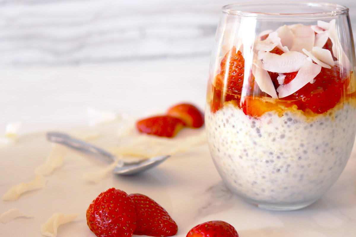 a clear glass layered with chia seed-quinoa pudding, honey, strawberries and coconut flakes on a marble counter top with a spoon and strawberry slices and coconut flaked scattered on top