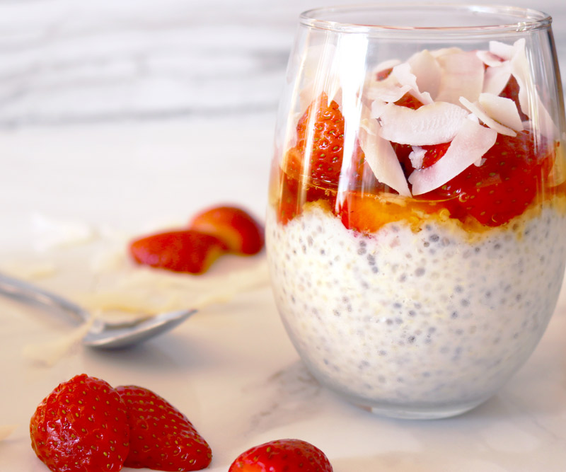 a clear glass layered with chia seed-quinoa pudding, honey, strawberries and coconut flakes on a marble counter top with a spoon and strawberry slices and coconut flaked scattered on top