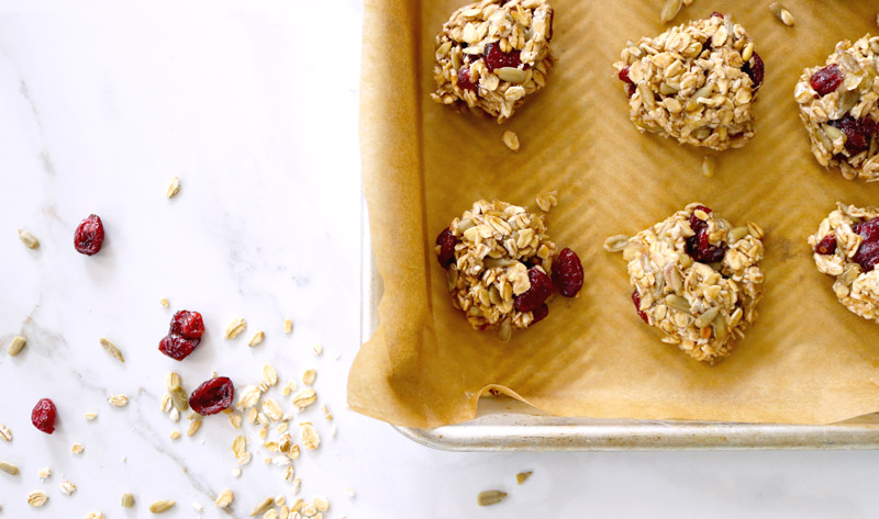 4-Ingredient Breakfast Cookies on a parchment-lined baking sheet sit on a marble counter top sprinkled with oats, sunflower seed kernels and dried cranberries
