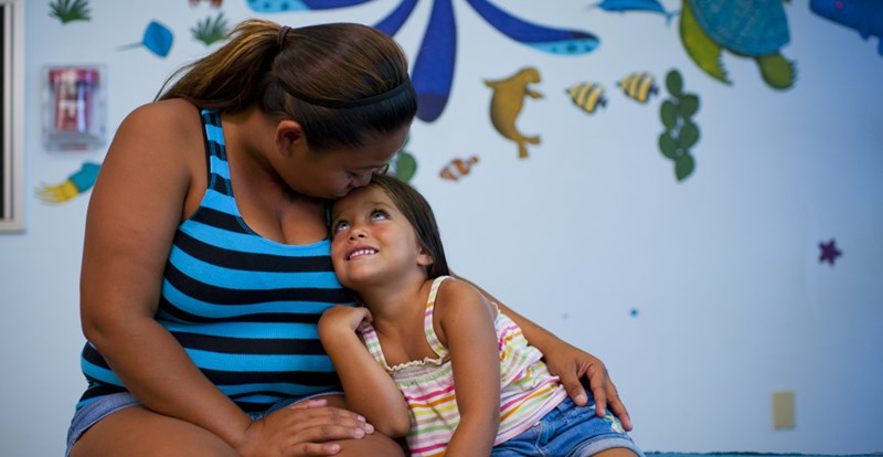 a daughter looks up at her mother with her mother kissing her on the forehead