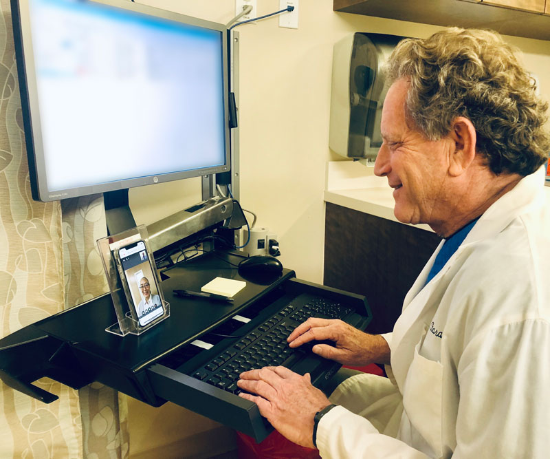 Dr. James Barahal sits at a computer and talks with a patient over a telehealth screen.