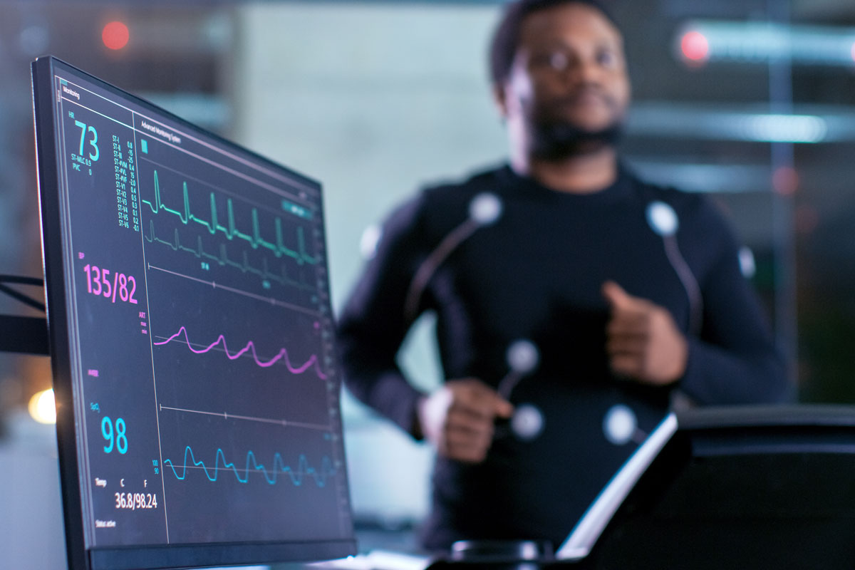 A man runs on a treadmill with sensors taped onto various parts of his body to track his heart rate as shown on a heart rate monitor.