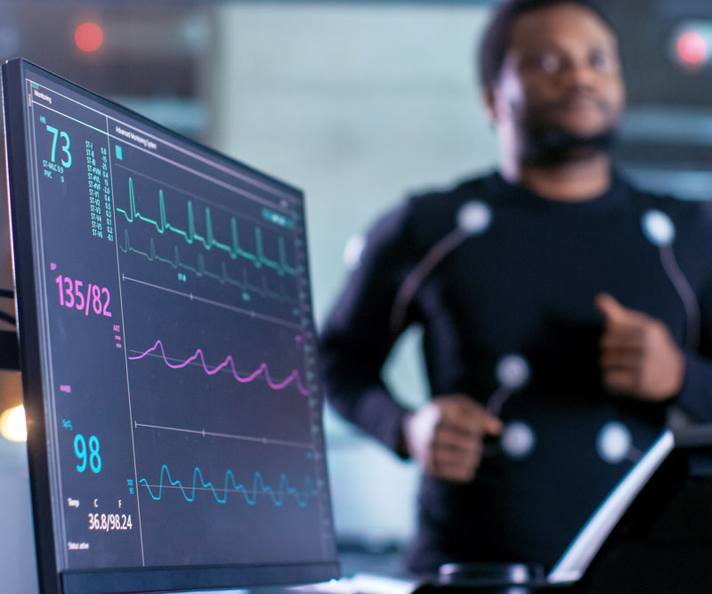 A man runs on a treadmill with sensors taped onto various parts of his body to track his heart rate as shown on a heart rate monitor.