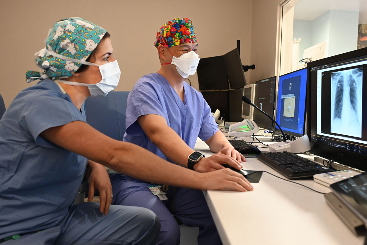 Two doctors dressed in scrubs look at a computer monitor in a children's hospital ward.