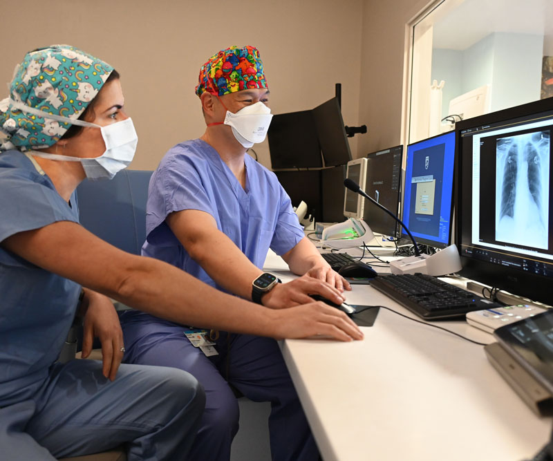 Two doctors dressed in scrubs look at a computer monitor in a children's hospital ward.