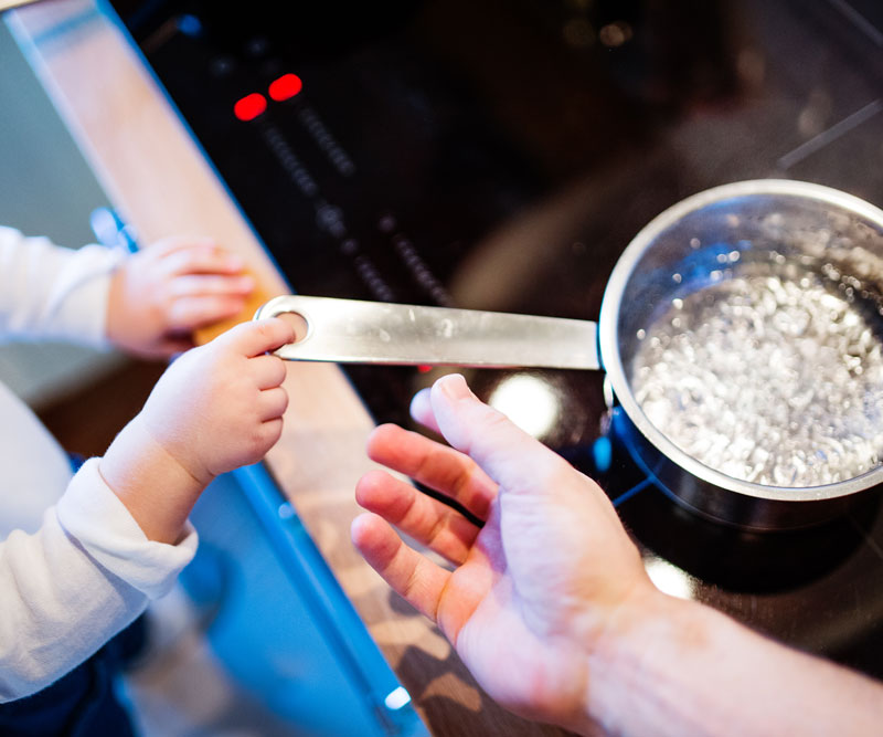 A young child's hands grab for the handle of a pot of boiling water as his parent's hands reach out to stop him.
