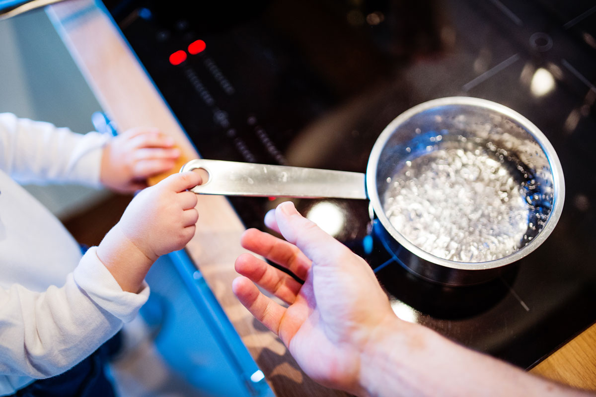 A young child's hands grab for the handle of a pot of boiling water as his parent's hands reach out to stop him.