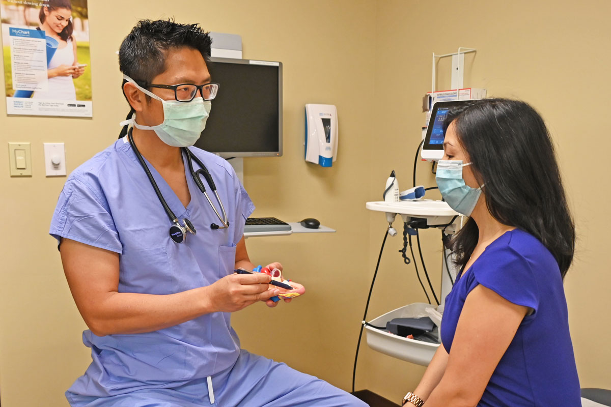 A physician wearing blue scrubs and a face mask sits in an exam room with a female patient and points to a model of a heart.