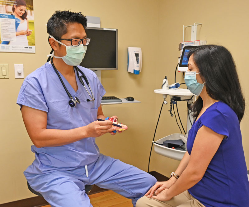 A physician wearing blue scrubs and a face mask sits in an exam room with a female patient and points to a model of a heart.