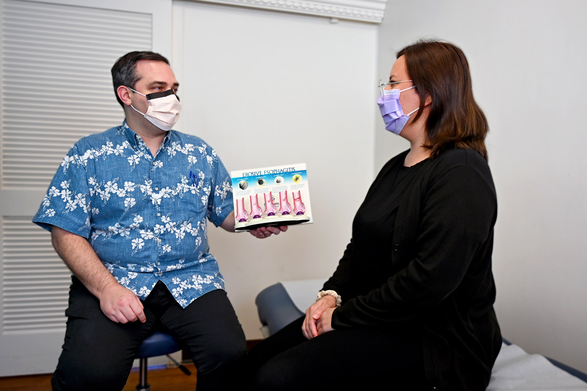 A male physician holds up a 3D model titled "Erosive Esophagitis" while talking to a female patient in an exam room.