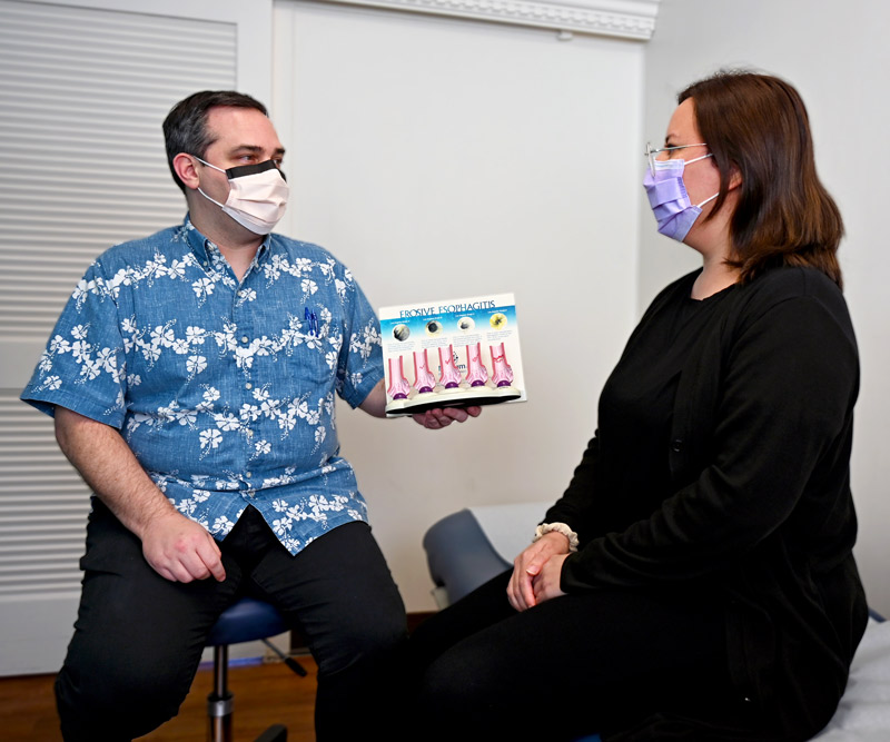 A male physician holds up a 3D model titled "Erosive Esophagitis" while talking to a female patient in an exam room.