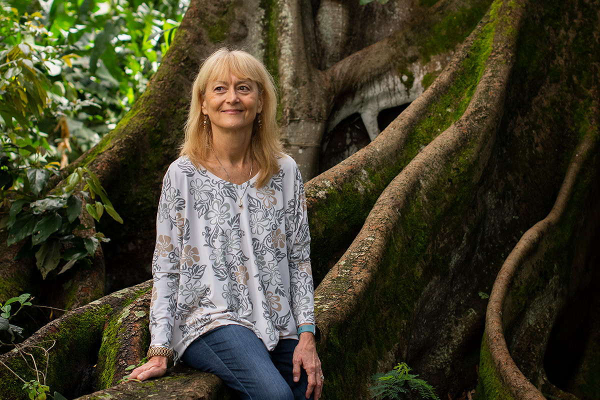 A woman sits on the roots of a banyan tree in a shaded forest.