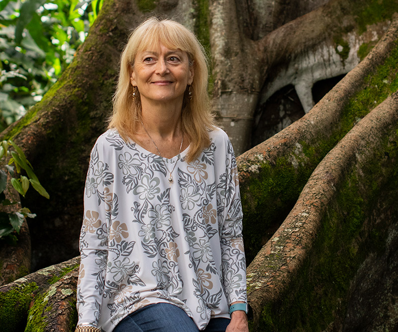 A woman sits on the roots of a banyan tree in a shaded forest.