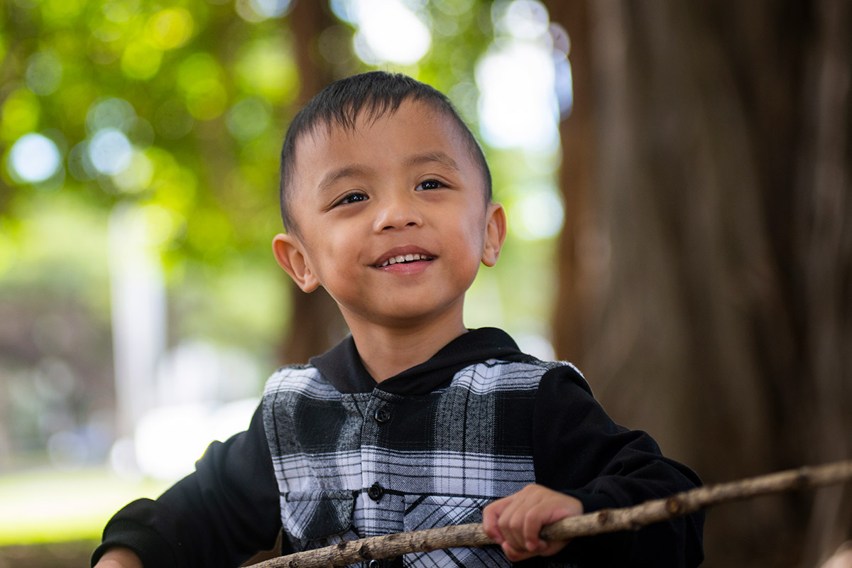 A young boy in a flannel jacket smiles at the camera as he stands in front of a blurred forest background.