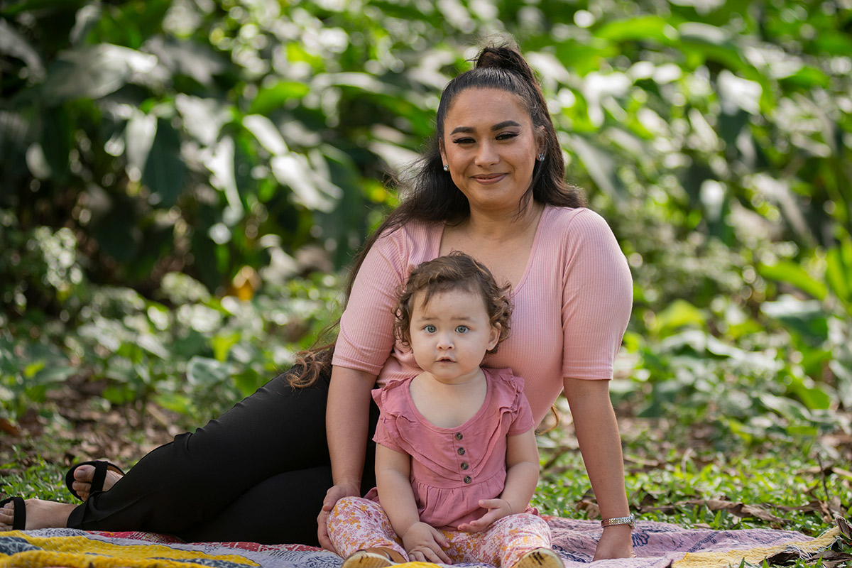 A mother and girl toddler sit on a colorful blanket surrounded by tropical plants.