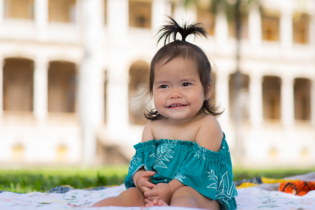 A 9-month-old girl sits on a colorful blanket in front of Iolani Palace and smiles at the camera.