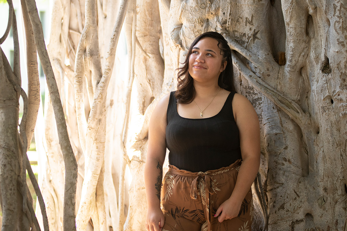 A teenage girl stands against the trunk of a large banyan tree and looks up peacefully at a patch of sunlight streaming through the branches.