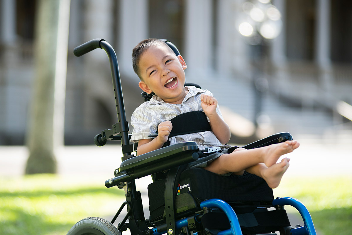 A young boy in a wheelchair smiles at the camera in front of Iolani Palace.