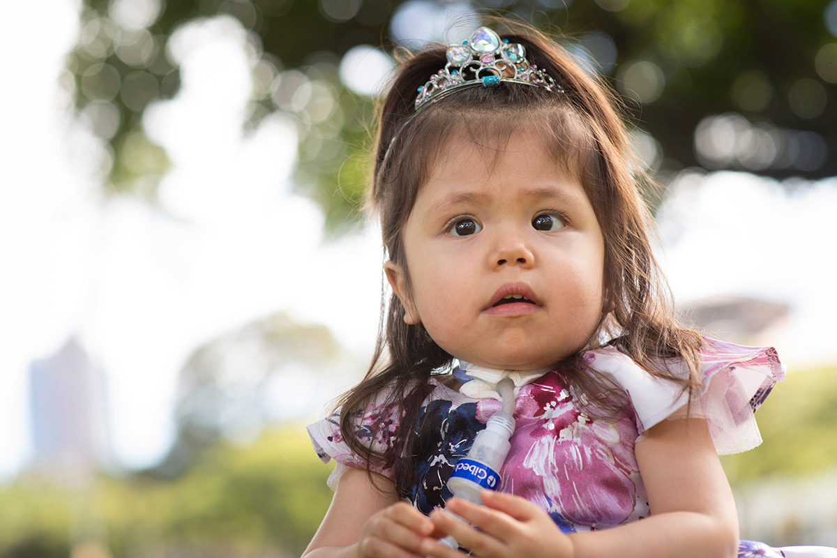 A 2-year-old girl with a breathing device is dressed as a princess and poses for the camera.