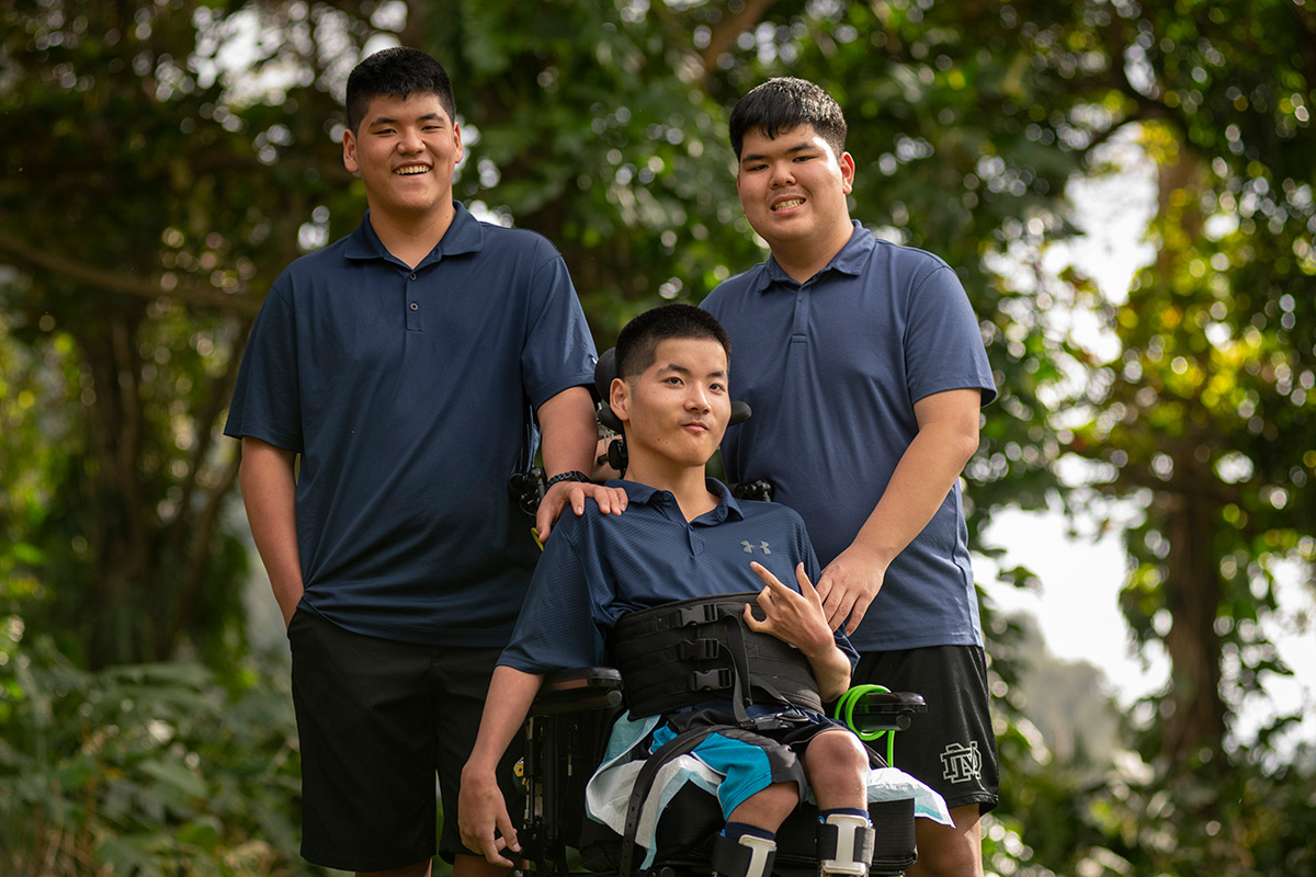 Two teenagers stand behind their triplet brother who is in a wheelchair against a forested background.