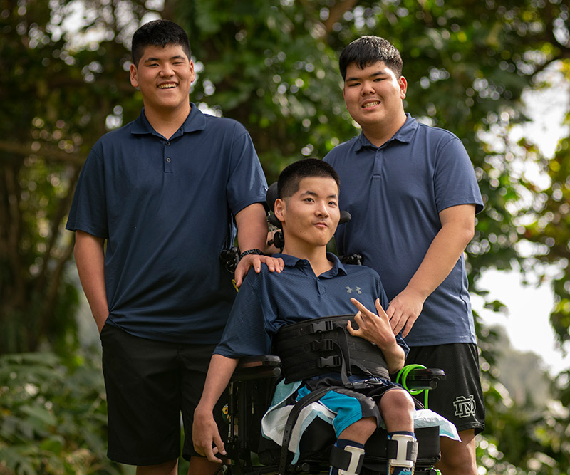 Two teenagers stand behind their triplet brother who is in a wheelchair against a forested background.