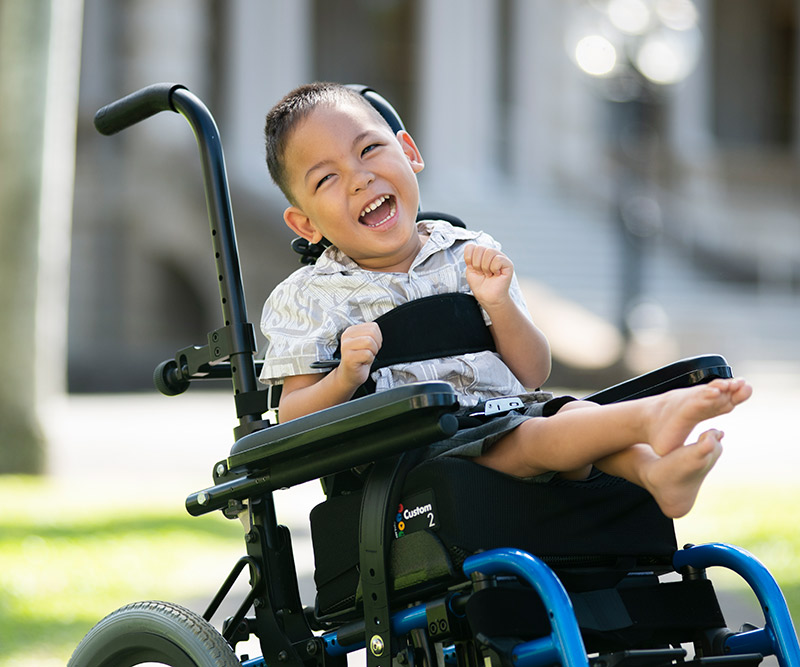 A young boy in a wheelchair smiles at the camera in front of Iolani Palace.