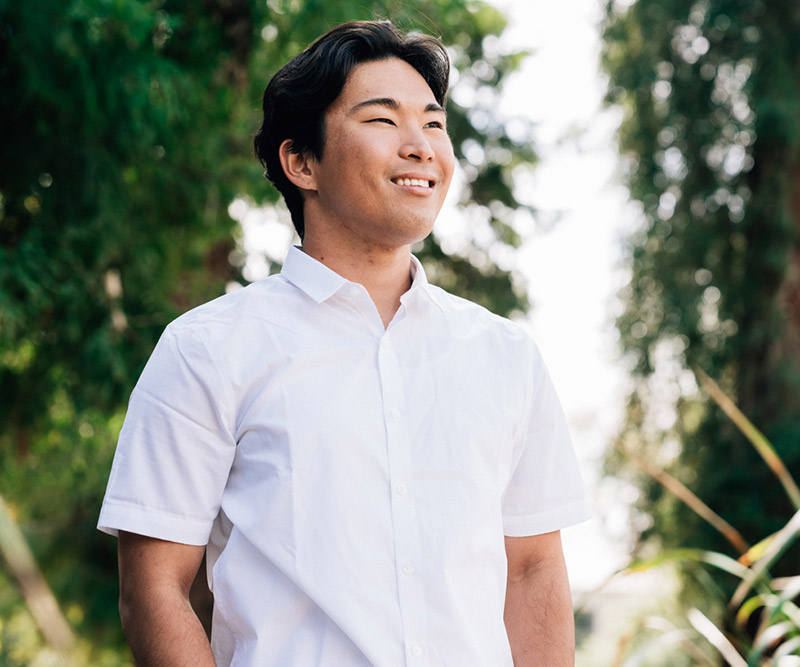 A young 20-somthing-year-old man stands looking hopefully out toward the distance against a forested background.