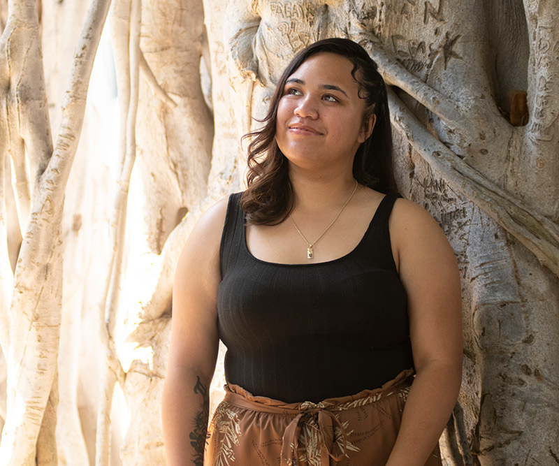 A teenage girl stands against the trunk of a large banyan tree and looks up peacefully at a patch of sunlight streaming through the branches.