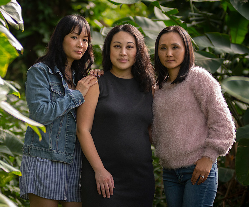 Three women stand closely together in a tropical rainforest.