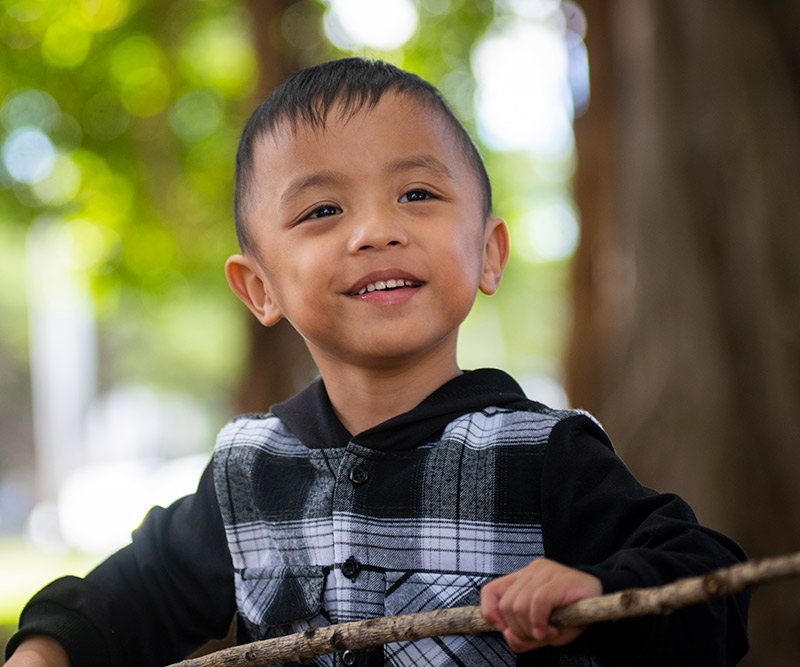 A young boy in a flannel jacket smiles at the camera as he stands in front of a blurred forest background.