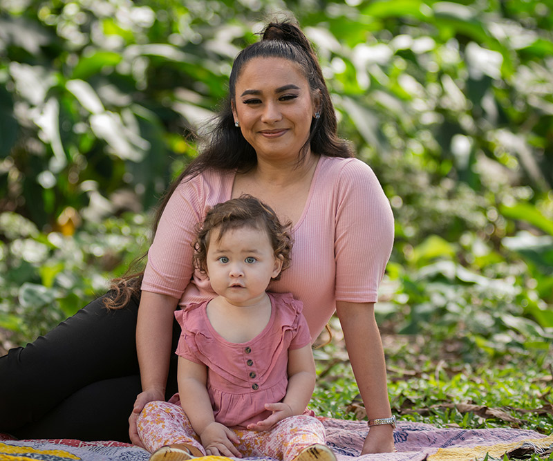 A mother and girl toddler sit on a colorful blanket surrounded by tropical plants.