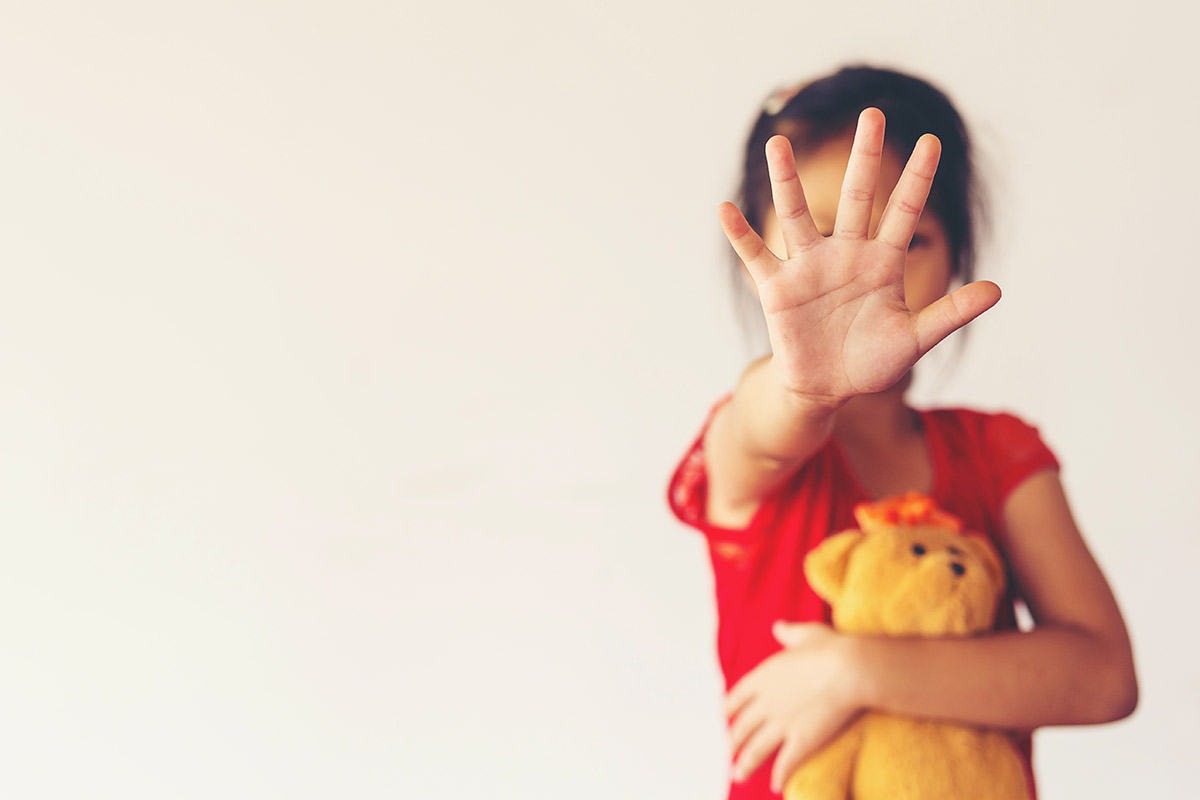 A young girl clutches a teddy bear in her right arm and holds her left hand up in front of her face to say "STOP!"