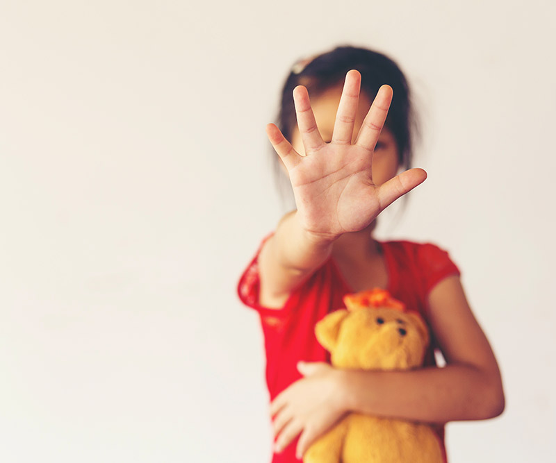 A young girl clutches a teddy bear in her right arm and holds her left hand up in front of her face to say "STOP!"