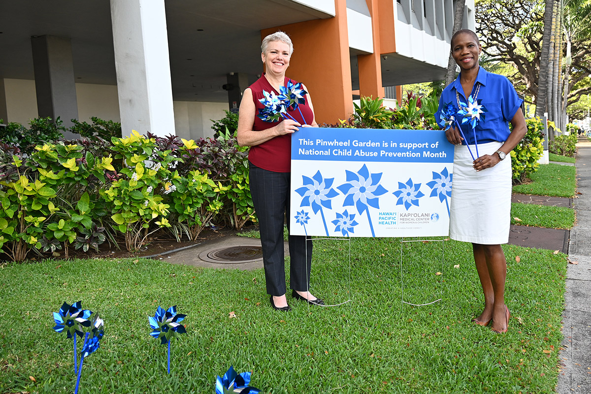 Two women stand on a grassy lawn in front of a children's hospital next to a sign that reads "The Pinwheel Garden is in support of National Child Abuse Prevention Month" and hold blue pinwheels.