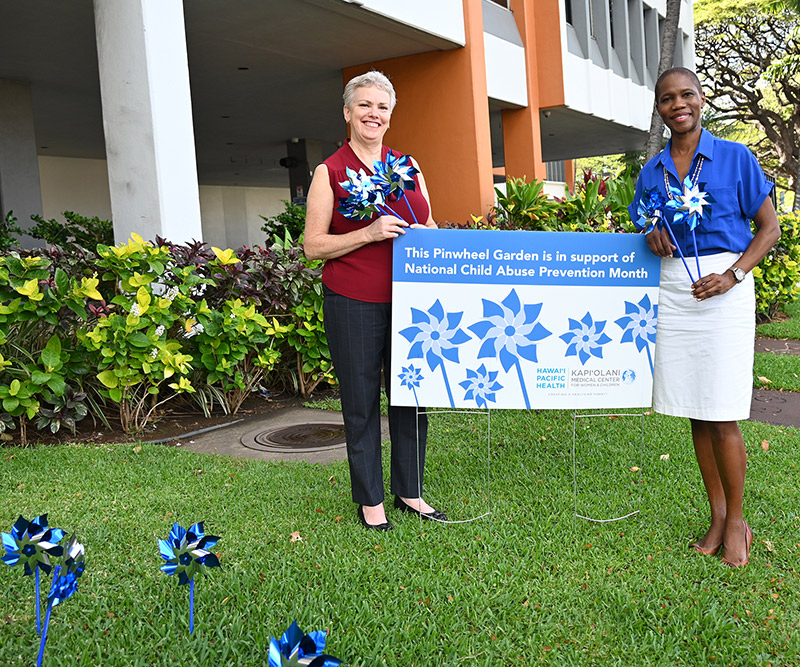 Two women stand on a grassy lawn in front of a children's hospital next to a sign that reads "The Pinwheel Garden is in support of National Child Abuse Prevention Month" and hold blue pinwheels.