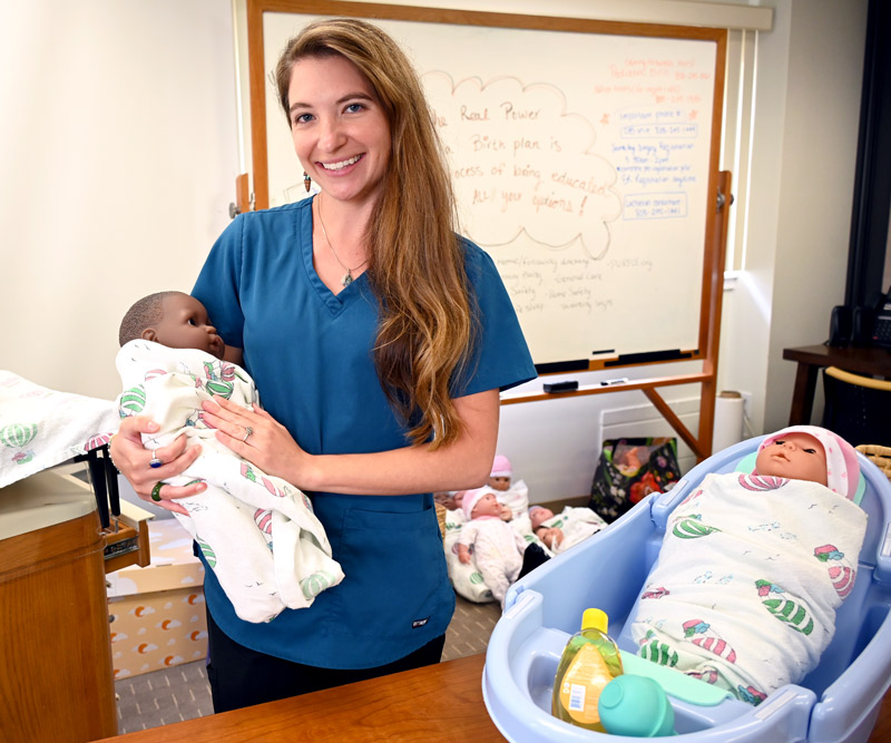 A woman wearing blue scrubs holds a model of a baby swaddled in a blanket in a childbirth education room at a hospital.