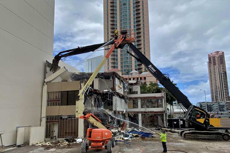 A ground level view of construction equipment and crew spraying down the dust from the demolition of the former Physical Therapy and Occupational Therapy Health Services building.