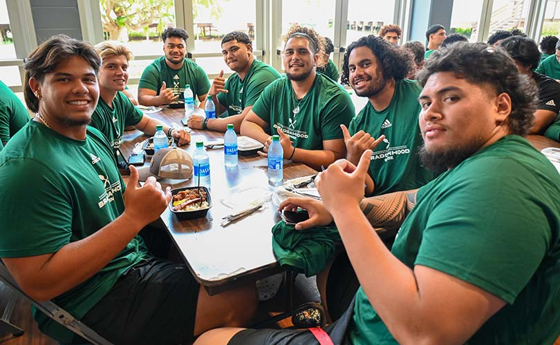 Seated group of Hawaii football players throwing shaka and smiling.