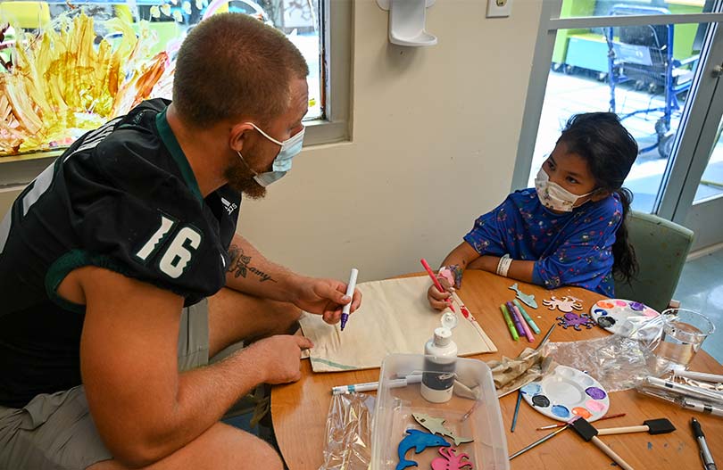 Football player sitting with young girl patient at small table coloring.