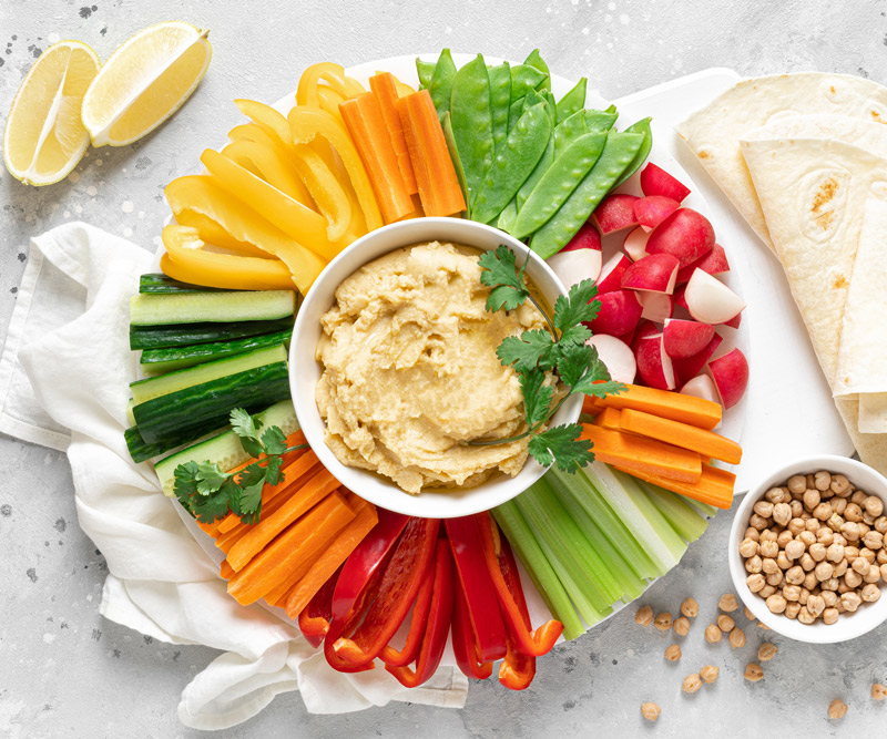a bowl of hummus surrounded by an assortment of fresh vegetable slices, garbanzo beans, lemon wedges and pita bread