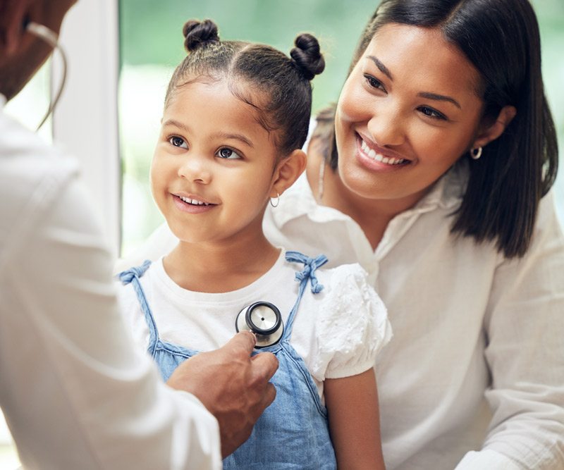 A doctor listens to a child's heartbeat using a stethoscope while her mother looks on with a smile.